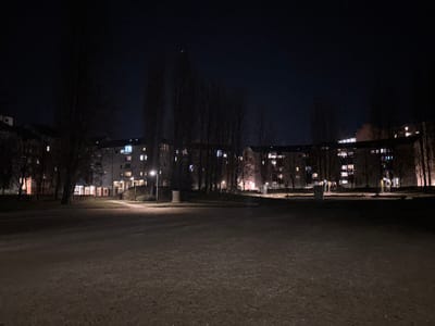 A night scene depicts a park-like area with a large grassy or dirt expanse in the foreground. Behind this expanse, a long, curved apartment building is visible, featuring numerous illuminated windows. Bare trees stand between the building and the open area, their branches silhouetted against the dark sky. A few lampposts provide sparse lighting along a pathway leading towards the building. The overall atmosphere is dark and quiet, with a sense of urban tranquility.