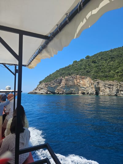 The image, captured from a boat traveling on the Adriatic Sea near Zagrađe, Montenegro, shows a picturesque coastal landscape. The foreground features the edge of the boat's roof, a white fabric awning supported by dark metal framing, with visible ropes and fixings. Below the awning, several passengers are seated, facing away from the camera, their backs and shoulders visible. They appear to be enjoying a boat tour.
The middle ground is dominated by the deep blue, rippling water of the sea, with white foam trailing behind the boat. In the distance, a dramatic cliff face rises, characterized by layered rock formations in shades of tan and gray, with patches of green vegetation clinging to its upper slopes. The cliff is densely covered in lush green trees and foliage, indicating a Mediterranean ecosystem. The sky is a clear, vibrant blue, suggesting a sunny day. The overall scene depicts a scenic excursion along the Montenegrin coast.
