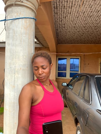 The image shows a woman lying on her side, with her head resting on a concrete pillar. She is wearing a pink tank top. Her eyes are closed and she appears to be sleeping or resting. The scene is outdoors, with a brick wall and a car visible in the background. The lighting suggests it is daytime. There is a blue rope tied around the pillar. A mobile phone is visible in the bottom right corner of the image. The setting appears to be in Russo, Nigeria.