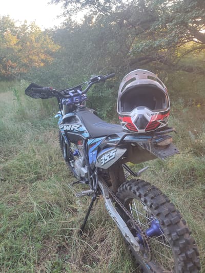 A dirt bike with a helmet resting on the rear fender is parked in a grassy area with trees in the background. The dirt bike is blue and black with the number "355" visible on the fender. The helmet is white, red, and black. The scene appears to be outdoors in a natural environment. There is no indication of the location being in the city of Novomar'evskaya, Russia.