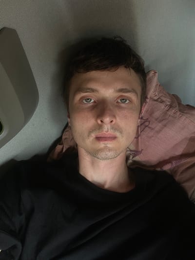 The media file shows a close-up, sideways shot of a young man lying on his back. He has fair skin, short brown hair, and light green eyes. He is wearing a black t-shirt. His face is partially illuminated, with some stubble on his chin and jawline. A pink and purple striped pillow is visible behind his head. The man appears to be in a relaxed or perhaps tired state, with a neutral expression. The background consists of a textured gray surface and a part of a gray object in the lower left corner, which could be part of a car interior or furniture. The lighting suggests it could be indoors, possibly during the day or evening. There is no visible text in the image. The location context provided is Brachterbeek, Netherlands.