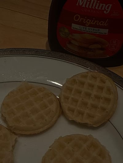 The image displays a close-up, top-down view of a breakfast meal in a dimly lit indoor setting. A white ceramic plate, featuring a dark, ornate rim, occupies the majority of the lower and central frame. Resting on the plate are at least three small, circular, light golden-brown waffles, each displaying a uniform grid pattern. In the upper right background, a dark bottle with a red label is partially visible. The label identifies the product as "Milling Company Original LITE SYRUP," with the "Aunt Jemima" brand prominently displayed. Further legible details on the label include "50 calories per serving," "12 FL OZ (355 ML)," and a "CHEFSBEST" seal. The plate and syrup bottle are positioned on a light brown wooden surface, likely a table or countertop. No specific location within Hallandale Beach, United States, is identifiable from the visual content.