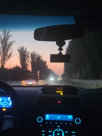 The image depicts the interior of a car driving on a road at dusk, with trees lining the right side of the road and a soft orange and blue gradient in the sky. The view is from the driver's perspective, showing the dashboard with illuminated displays. On the left side of the dashboard, a digital speedometer shows a speed of 93 and another number, 66716. The center console features a radio display with illuminated blue buttons and a circular control knob. A small red triangle warning light is visible on the dashboard. In the distance, headlights of other vehicles can be seen on the road, and a truck with illuminated taillights is ahead. The rearview mirror hangs from the windshield, and a dashcam is mounted below it. The location is identified as Sariqmirzachek, Uzbekistan.