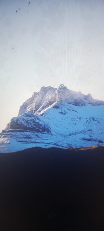 The image shows a snow-covered mountain range against a bright sky. The mountain has sharp peaks and slopes covered in snow and ice. The lower part of the image is a dark silhouette, likely the foreground. There are several small dark specks visible in the sky, possibly dust or imperfections on the camera lens. There are no people, animals, or man-made structures visible in this image. The location in Kudymkar, Russia is not discernible from the visual information present.