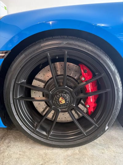 A close-up, low-angle shot shows a bright blue Porsche car's front wheel. The wheel is black with a multi-spoke design. A large red brake caliper with the word "PORSCHE" printed in white is visible. The brake rotor has numerous small holes. The tire is black and has text on its sidewall. The car is parked on a concrete surface. The background consists of a white wall and a section of the car's blue fender. The location is Wisconsin Dells, United States of America.