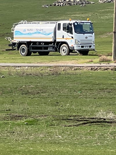 The image displays a white tanker truck situated on an unpaved dirt path, next to a vast green field under bright daylight. The scene suggests an outdoor, possibly rural or agricultural setting.
The main subject is a medium-duty truck, painted white. Its cab features a prominent silver grille with a circular emblem in the center, and a black bumper. Amber lights are visible on the roof of the cab, possibly clearance lights or turn signals. Above the windshield, a sun visor or decal reads "NURLI TO'LKIN" in black capital letters.
Attached to the truck's chassis is a white cylindrical tanker body. This tanker is adorned with a graphic design featuring a light blue wave pattern and an orange stripe. On the side of the tanker, the words "NURLI TO'LKIN" are printed in dark blue capital letters, accompanied by two stylized blue musical notes or water droplets to the left of the text. The truck has visible tires with black rims, and a dark-colored chassis and frame.
To the immediate right of the truck, a narrow, light-colored dirt path or road runs parallel to it. Beyond this path, a wide expanse of bright green field stretches into the background. The field shows some variation in texture and color, with hints of drier vegetation or soil mixed with the lush green.
In the far left background, a pile of rocks or rubble is visible. A wooden post or fence pole stands upright on the far left edge of the image. The strong, clear lighting indicates a sunny day. No people are visible in the image.