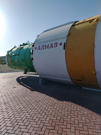 This is a photograph of the Almaz space station, displayed outdoors on a sunny day. The Almaz is a large, cylindrical object, primarily painted white with yellow and green sections. Red lettering spelling "АЛМАЗ" (Almaz) is prominently displayed on the white section, accompanied by a red star symbol. The object appears to be a decommissioned space module, possibly part of a museum exhibit.
The Almaz is situated on a patterned brick or paver walkway, which is partially shaded by the object itself. In the background, a building with a light-colored facade can be seen, along with a few sparse trees and a person walking in the distance. The sky is clear and blue, indicating fair weather. The sun is casting distinct shadows, suggesting it is either morning or late afternoon. The overall impression is one of a static display in an open-air setting, likely a public space or park, given its exhibition. The context of Baikonur, Kazakhstan, suggests this might be related to the history of Soviet and Russian space exploration. No specific activity is taking place; the image captures a still moment of the Almaz space station on display.