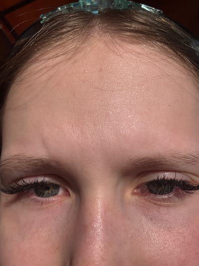 The image shows a close-up of a person's face, focusing on their eyes, forehead, and hair. The person has light skin, dark eyelashes, and gray eyes. A small portion of a jeweled headband is visible in their hair. The background is dark and out of focus. The city of Lunezhki, Russia is not visible or apparent from the given image.