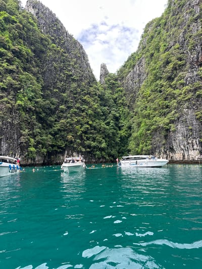 The image shows a scenic view of a rocky, jungle-covered cliff face rising from a calm, turquoise body of water. Several white boats are docked near the shore. One boat, labeled "Diamond Sea," has people on board. Another boat, further in the foreground, has "SUZEAN" written on its side. The water is clear and reflects the sunlight, creating shimmering patterns. Small buoys with yellow tops are visible in the water, suggesting a designated swimming or docking area. The sky is partly cloudy, and the overall lighting suggests it is daytime. The lush greenery on the cliffs indicates a tropical environment. The scene evokes a sense of relaxation and leisure, typical of a tropical vacation destination like Haad Yao, Thailand.