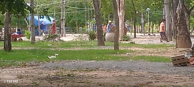A daytime scene in Thmar Puok, Cambodia, shows a partially cleared area with trees and patchy grass. In the mid-ground, several individuals are present. To the left, a child sits in a red wagon near a blue tarp structure. Further right, a person in a red garment stands facing left. In the center, a person wearing a straw hat and a blue shirt is near a large ceramic pot. To the far right, a person in an orange top and patterned skirt stands facing left. The ground is covered with gravel and scattered debris, including a pile of bricks in the lower right corner. The timestamp "2026/06/18 09:16" and "©TECNO" are visible in the bottom left corner.