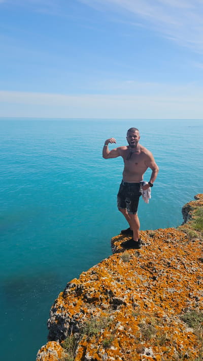 A man stands on the edge of a rocky, moss-covered cliff overlooking the turquoise sea at Kamen Bryag, Bulgaria. He is shirtless, wearing dark shorts, and flexing his bicep while shouting. He holds a white towel in his hand. The sky is clear blue.