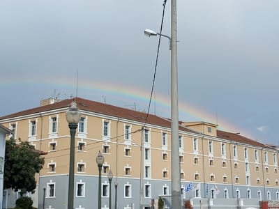 The image depicts a three-story building with a red tile roof in the city of Drama, Greece. The building's exterior features a two-tone color scheme, with a tan upper section and a gray lower section. The building has numerous windows, and several flags are visible near the entrance. Street lamps line the sidewalk in front of the building. In the background, a rainbow arcs across the sky. Overhead power lines and a utility pole are visible in the foreground. A tree is visible on the left side of the frame.