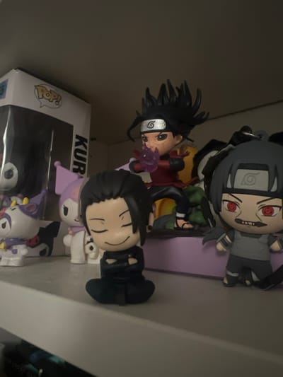 A shelf contains anime figurines. At the top is a keychain of Itachi Uchiha, a figure of Sasuke Uchiha performing a jutsu, and a figure with dark hair. Below them are a Funko Pop box with "KUR" visible, and several figurines of a character that resembles Kuromi. The lighting is dim.