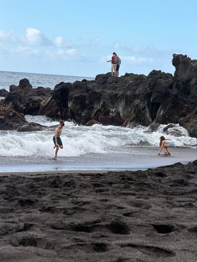 This media file depicts a scene at a rocky beach in Puerto de la Cruz, Spain. There are at least three people present. In the foreground, a young man is standing on a wet, sandy shore, facing away from the camera, with waves breaking around his feet. He appears to be wearing swim trunks. Further back and to the left, three individuals are standing on dark, rocky terrain overlooking the ocean. Two of them are wearing backpacks, suggesting they may be hikers or tourists. Another person, a young woman, is kneeling on the wet sand closer to the water, with her arms extended.
The scene is outdoors, with a predominantly grey sky, indicating overcast weather. The time of day appears to be daytime. The environment is characterized by dark, volcanic-like rocks and dark sand, typical of some coastal areas in the Canary Islands. The ocean is visible, with noticeable wave activity. The overall mood is relaxed and recreational, with people engaging with the natural environment. There is no visible text in the media file.
