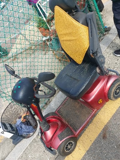 A dark red four-wheeled mobility scooter is positioned on a light gray concrete curb adjacent to an asphalt surface with two parallel yellow painted lines. The scooter features a black seat with a bright golden-yellow backrest cushion, black handlebars, and two black rear-view mirrors. A black wire basket attached to the front contains a blue fabric item. The scooter's body shows signs of use and minor dirt accumulation. In the background, a green chain-link fence separates the curb from an area containing several potted plants, including green foliage and a terracotta pot. The lower legs and feet of an individual, wearing dark pants and black athletic shoes with white soles, are visible on the right side of the frame, standing on the asphalt near the scooter. The scene is outdoors and well-lit. The location is 강화군, South Korea.