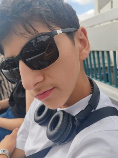 The image presents a close-up, slightly angled portrait of a young individual, likely male, with dark hair, prominent nose, and a faint mustache. He wears black square-frame sunglasses featuring a silver accent on the temples. A white crew-neck t-shirt is visible, and black over-ear headphones are worn around his neck, resting on his chest. A silver-toned metallic link watch is present on his left wrist, and a dark strap, possibly from a bag or seatbelt, diagonally crosses his chest.
In the background, to the left, a portion of another individual wearing a dark top and blue jeans is visible, suggesting the subject is seated next to someone. To the right, a light-colored, possibly white, structure or railing is present, with vertical greenish-blue bars behind it. The sky, showing a mix of blue and white clouds, is visible above this structure.
The reflection in the sunglasses provides critical contextual details: railway tracks or a road with a white line or curb, and vertical architectural elements consistent with a platform or overhead structure. A human hand holding a smartphone, indicative of the photographer, is also clearly reflected in the right lens. The scene depicts an individual seated in an outdoor or semi-outdoor setting consistent with a transport hub. The environment, including railway infrastructure and elevated structures, indicates the location is the RER station in Neuilly-Plaisance, France.