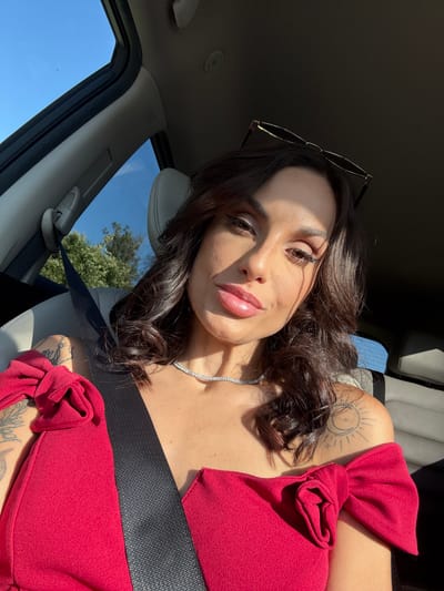 A woman with medium-length brown hair, wearing a red top and a silver necklace, is seated in a car. She is wearing a black seatbelt. Sunglasses are perched on her head. She has tattoos on her shoulder and arm. The car interior is light-colored. Trees and blue sky are visible through the window. The location is unknown, the information about Kaleytsa, Bulgaria is incorrect.