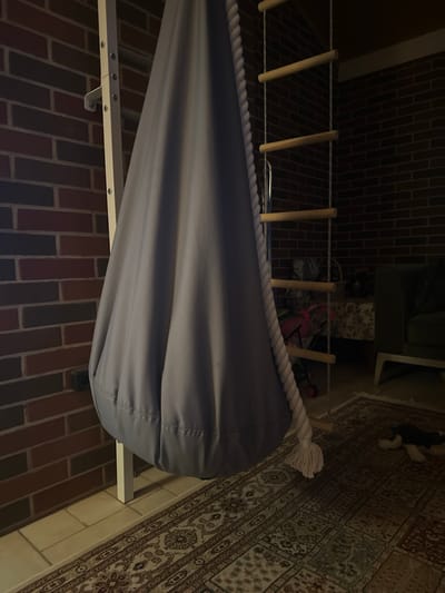 The image shows a gray fabric swing hanging from a wooden ladder that is attached to a brick wall. The ladder is on the left side of the frame, and the swing occupies the central portion. The swing is made of a thick, textured fabric and has a rope handle. The background shows a dimly lit room with a rug on the floor and a couch in the distance. The brick wall is a prominent feature, with dark red and brown bricks. The lighting suggests it is evening or night. The scene appears to be indoors, possibly a child's playroom or a recreational area. There are no people visible in the image.