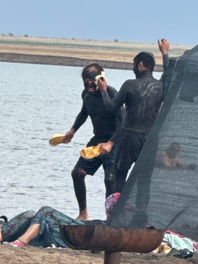 Two men covered in mud are by the water's edge in Ямышево ауылдық округі, Kazakhstan. The man on the left, with his face covered in mud, holds a yellow sandal in his right hand. He is wearing a black long-sleeved shirt and shorts. The man on the right, also covered in mud, is seen from behind, with his left arm raised. He is wearing a black long-sleeved top and dark shorts. In the foreground, a person is lying down on the sand, covered by a blue patterned cloth. Another person wearing a colorful headscarf is partially visible behind the lying person. A dark-colored fabric structure is present on the right side of the image, partially obscuring the scene. The background shows a body of water and a distant, dry, grassy landscape under a hazy sky.