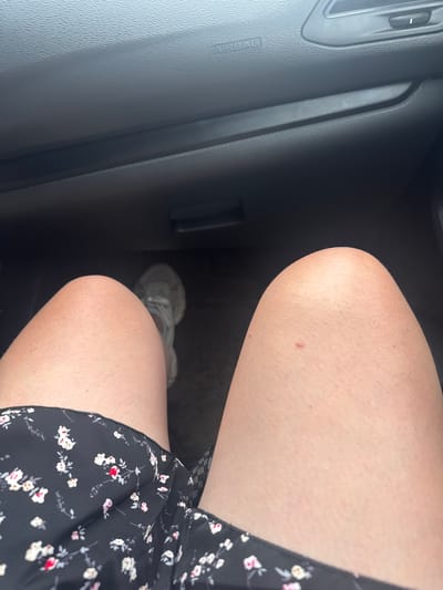 The image shows a person's legs and a portion of a car's interior. The person is wearing a black floral dress and white sneakers. The car interior includes the dashboard with an "AIRBAG" label and an air vent. The lighting suggests it is daytime. The location is specified as the city of Permyakovka, Russia.