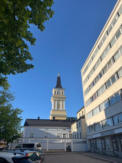The image captures a street scene in Oulu, Finland, on a clear, bright day. Dominating the right side of the frame is a modern, multi-story building with a light-colored facade and numerous rectangular windows. To the left, a classic yellow and white church tower with a black spire and a clock face stands prominently against the vivid blue sky. In the foreground, a section of a light blue, traditional wooden building with a dark roof is visible, bordered by a white fence. Several cars are parked on a cobblestone street in front of the buildings. Green foliage from a tree hangs into the upper left corner of the image, framing the sky. Shop signs, including "BLONDIA" and "HUUSEN MINNAK," are visible on the ground floor of the modern building. The overall impression is one of an urban environment that blends historical and contemporary architecture.