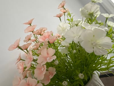 The image displays a close-up view of a floral arrangement, predominantly featuring delicate pink and white artificial flowers. The flowers are clustered together with vibrant green artificial foliage. The arrangement is placed against a plain, light-colored background that appears to be a wall. There is no discernible presence of people or any specific location within Kattakurgan City, Uzbekistan, identifiable from the visual information presented. The focus is solely on the artificial flowers and greenery, creating a simple and decorative composition.