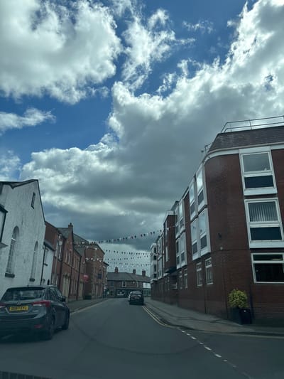The image shows a street scene in Knutsford, United Kingdom. The view is of a street lined with buildings of varying heights and architectural styles, a mix of brick and white-painted facades. Bunting is strung across the street. The sky is filled with both blue sky with scattered white clouds and a large mass of grey storm clouds. A dark colored vehicle is parked on the right side of the street in the foreground. Another dark colored vehicle is further down the street. The street is paved and has painted yellow lines denoting parking areas.
