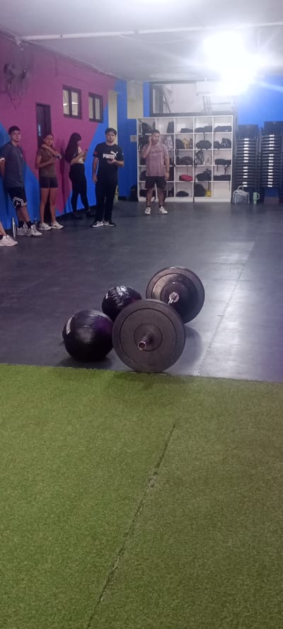 An interior view of a fitness facility located in Quilmes, Argentina, shows five individuals and various gym equipment. In the foreground, a section of green artificial turf occupies the lower left corner, meeting a dark grey rubber mat floor. On this mat, four pieces of equipment are visible: two black medicine balls of different sizes and two black bumper plates resting on a barbell, with the barbell shaft extended between them.
In the mid-ground, five individuals are standing. From left to right: a male in a dark t-shirt and shorts stands partially obscured; next to him, a female in a dark top and shorts looks at a smartphone; another female in a light crop top and dark leggings also looks at a smartphone, her back partially turned; a male in a black PUMA t-shirt holds and looks at a smartphone; and furthest right, a male in a light brown t-shirt and dark shorts stands with his left hand near his left ear.
The background features a wall painted with geometric sections of pink, blue, and purple. Two dark-framed windows are visible on the pink section, along with a wall-mounted fan. To the right, a white shelving unit with multiple compartments holds various bags and items. Further right, a tall stack of dark chairs or stools is positioned against a blue wall. Overhead lighting sources illuminate the scene, with a prominent glare originating from the upper right portion of the ceiling.