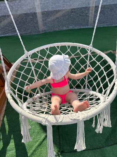 A baby is lying in a white macrame hammock chair. The baby is wearing a white sun hat and a pink bikini. The hammock is suspended from above by ropes and is hanging over a green, artificial turf surface. In the background, there is a blurred area that appears to be a gray tarp or sheet, possibly covering something. The scene is outdoors, and the bright sunlight suggests it is daytime, likely a warm day given the baby's attire. The overall impression is one of a relaxed, leisurely moment, perhaps at a backyard or resort setting. The baby's posture and the pacifier in its hand suggest a moment of rest or play. There is no visible text in the image.