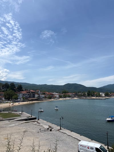 This image captures a scenic view of the coastal town of Olympiada, Greece. The foreground features a concrete embankment with a white van bearing "MERCURY MerCruiser" and "TWIN DISC" logos parked to the right. Further left on the embankment are some scattered pieces of lumber and a trailer. Beyond the embankment, a body of calm, dark blue water is visible, with several small boats and a larger white and blue boat anchored.
The middle ground reveals a sandy beach populated with sunbathers and umbrellas. Behind the beach, a row of buildings with red-tiled roofs and white facades lines the shore. These buildings appear to be hotels or restaurants. A long wooden pier extends from the beach into the water.
The background is dominated by lush green, rolling hills that rise steeply behind the town. The sky above is a bright blue with scattered white clouds, indicating fair weather. The overall impression is one of a peaceful seaside resort on a sunny day.