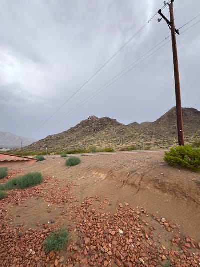 The image depicts a rural scene with mountains in the background under a cloudy sky. A wooden utility pole with power lines stands prominently on the right side of the frame. The foreground is a mix of reddish-brown gravel and sparse green vegetation. A road is visible leading towards the mountains. Part of a red-tiled roof is visible on the far left. The location is identified as Etholen, United States. There are no people or animals visible in the image. The environment suggests a dry, arid climate.