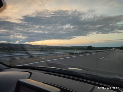 An eye-level, wide shot captures a sunset viewed from the interior of a vehicle on a highway. The sky is filled with a dramatic display of dark gray and white clouds, with a sliver of orange sunlight peeking through the horizon. The highway stretches into the distance, lined with metal guardrails and a white dividing line. The landscape beyond the highway is hilly and covered with greenery. The dashboard of the car is visible in the foreground, and a date and time stamp "13.06.2026 05:51" is present in the bottom right corner. The scene is set in Kalugerovo, Bulgaria.