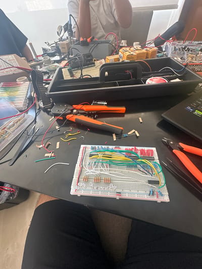 The image depicts a cluttered workspace, likely indoors, with a focus on electronics projects. In the foreground, there is a breadboard with several electronic components, including resistors, LEDs, and integrated circuits, connected by wires. Wire strippers and wire clippings are also present on the workspace. There is a black laptop, likely used for programming. Boxes of electronic components and other equipment are visible. There are two people present, one has their face blocked from view, but their arm is visible. The location is not explicitly stated and cannot be determined from visual clues.