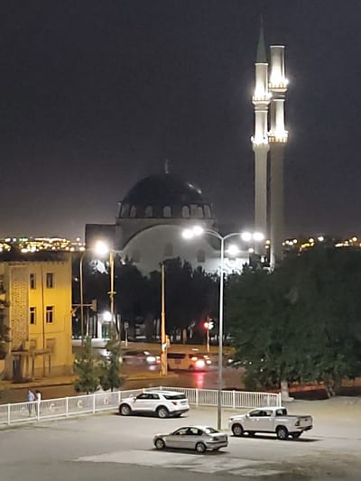 The image depicts an outdoor scene at night, likely in Ashgabat, Turkmenistan. The primary subjects are architectural structures and several cars. Dominating the left side of the frame is a minaret of a mosque, illuminated by lights. The minaret is tall and slender, with decorative elements and a pointed spire. Adjacent to it, another, shorter minaret is visible. In the background, a large, dark dome, characteristic of mosque architecture, is partially obscured by trees.
To the right of the minarets, a row of white cars is parked along a white railing, which appears to be a pedestrian walkway or barrier. The cars are modern sedans. Beyond the cars, the lights of what seems to be a city are visible in the distance. There are also streetlights and traffic lights, suggesting an urban or semi-urban environment. The overall lighting suggests it is nighttime, with artificial lights illuminating the mosque and surrounding areas. The weather appears to be clear.
The scene is static, with no obvious activity taking place other than the presence of the parked cars and the illuminated buildings. The overall mood is serene and perhaps even imposing due to the scale of the mosque architecture against the night sky. There is no visible text in the image. The location context of Ashgabat, Turkmenistan, aligns with the prominent mosque architecture.