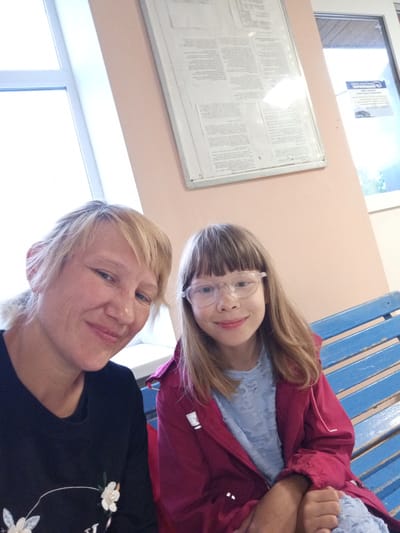 Two individuals, an adult and a child, are captured in a selfie, likely taken indoors. The adult, with blonde hair and fair skin, is positioned in the foreground on the left, smiling. The child, with light brown hair and wearing glasses, is seated to the right, also smiling. They are both wearing casual clothing. Behind them, a blue slatted bench is visible, suggesting they are in a waiting area or public space. The background includes a pale pink wall with a bulletin board containing text and images, and a window with a view of the outdoors. Based on the provided information, this scene is located in the city of Verkhneyarkeyevo, Russia.