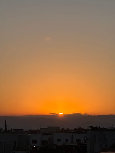 This is a landscape photograph taken at sunset in Katzrin, Israel. The image is oriented vertically, with the sun setting behind a mountain range on the right side of the frame. The sky is filled with a gradient of warm colors, transitioning from a pale yellow near the horizon to a muted orange and then a soft gray at the top of the image. A thin strip of clouds is visible just above the mountain peaks, catching the last rays of the sun.
In the foreground, silhouetted against the bright sky, are the rooftops of buildings. These appear to be residential structures, with distinct architectural features like windows and balconies. Trees and a tall, thin cypress tree are also visible in silhouette, adding to the urban or suburban setting. Streetlights can be seen on poles among the buildings, some of which are illuminated, suggesting the transition from daylight to evening.
The overall mood of the image is peaceful and serene, characteristic of a sunset. The lighting indicates it is late evening, shortly before or during twilight. There is no visible text in the image. The presence of buildings and what appears to be a developed landscape indicates an inhabited area.
