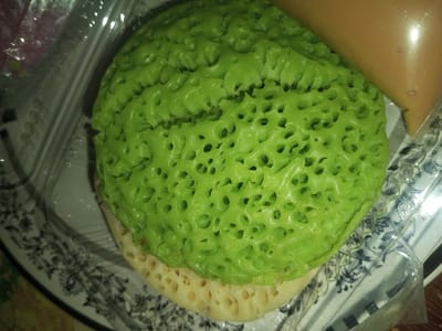 A close-up, overhead photograph displays a green and cream-colored food item contained within a clear plastic takeaway box, set upon a patterned plate. The prominent upper layer of the food is bright green, exhibiting a highly porous, spongy texture with numerous irregularly sized holes. A cream-colored, similarly textured layer is visible beneath the green portion. A translucent pouch containing a light brown, viscous liquid is positioned alongside the food within the plastic box. The plastic box shows reflections of ambient light. The underlying plate is white with dark, ornate floral designs. This scene is located in Doramamidi, India. No individuals are visible, and no actions or interactions are depicted.