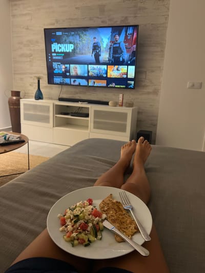 This image depicts an indoor living space, likely an apartment or hotel room, located in Almancil, Portugal. A person is observed from a first-person perspective, lying on a grey upholstered bed or sofa. Their bare lower legs and feet are visible, extended towards the television. On their lap rests a white round plate containing a meal, consisting of a mixed salad with what appears to be cucumber, tomato, and feta cheese, alongside two pieces of grilled poultry, likely chicken. A silver fork and knife are placed on the plate.

In the background, a large flat-screen television is mounted on a wall featuring light-colored horizontal paneling. The TV screen displays the Amazon Prime Video interface, with "PICKUP" highlighted as a primary title, showing two male figures in tactical vests and a red vehicle in the background. Below the TV is a white media console with multiple compartments, some with frosted glass doors. A black soundbar is positioned on top of the console. To the left of the console, a tall, dark brown terracotta-style urn stands on the floor. On the console, various small decorative items are present, including a blue vase with a feathery plant, two small light-colored spherical objects, and a rectangular container. To the left of the bed, a round wooden coffee table with a metal base is visible, holding a magazine or book on its surface and resting on a textured, light brown rug. The overall environment is well-lit and appears to be a contemporary setting.