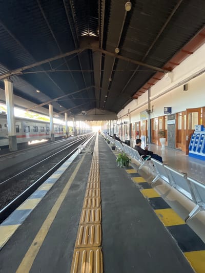 The image depicts a train station platform in Desa Tambakreja, Indonesia. The platform is covered by a dark corrugated metal roof supported by white pillars. Two train tracks run parallel along the platform. On the left side, a white and red train is stopped at the platform. On the right side, several rows of empty silver benches are arranged for waiting passengers. Some potted plants are placed near the benches.
Further down the platform, a few people are seated on the benches, some looking at their phones. A charging station with multiple ports is visible on the right side of the platform. The walls of the station building are painted white and feature several wooden doors and windows. Signs are mounted on the wall, including one with an "i" symbol and text that appears to be in Indonesian. The ground of the platform is paved with dark asphalt, marked with yellow and black lines for safety. A tactile paving strip is present along the edge of the platform. Sunlight streams in from the end of the platform, creating a bright, elongated perspective.