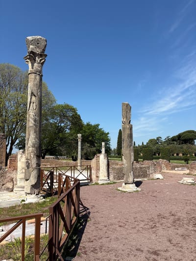 The image depicts ruins with ancient columns at Hadrian's Villa in Tivoli, Italy. Four stone columns stand somewhat intact, with fragments and capitals visible. A modern, brown metal fence surrounds portions of the ruins. The ground is covered in reddish-brown gravel. Trees and green vegetation are scattered throughout the site. A clear blue sky with wispy clouds forms the backdrop.