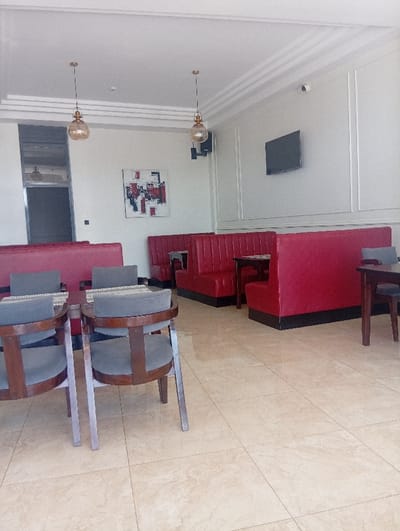 The image displays the interior of an empty dining establishment located in Murang'a, Kenya. The floor is surfaced with light beige rectangular tiles. The walls are painted a light, off-white color; the right wall features decorative vertical paneling, while the left wall displays an abstract painting composed of red, black, and white elements. A black flat-screen television is mounted on the right wall, and a white CCTV camera is affixed to the ceiling in the upper right quadrant. Two spherical, clear-amber glass pendant lights hang from the white ceiling, which also features crown molding. A black rectangular speaker is mounted on the left wall below one of the pendant lights. Seating arrangements include multiple red upholstered booth seats with black bases, accompanied by dark-stained wooden tables. In the foreground, several dark-stained wooden chairs with dark grey upholstered seats and backs are positioned around tables, one of which has a patterned placemat. A dark vertical opening, appearing to be a doorway or window, is visible on the far left. No individuals, actions, or interactions are depicted.