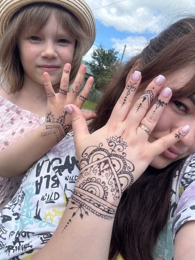 In Маркидоновка, Russia, a young girl wearing a straw hat and a pink dress poses with an adult woman. Both individuals have intricate black henna designs on their hands. The girl's henna is on her fingers, while the woman's henna extends from her palm up her arm. They are outdoors, as indicated by the blue sky with clouds and green trees in the background. The woman is wearing a multi-colored t-shirt. The scene appears to be a casual, outdoor gathering.
