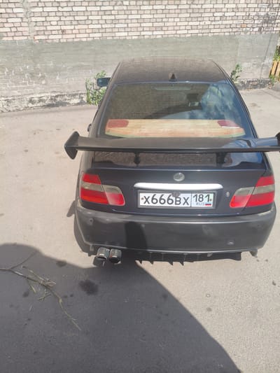 This media file shows the rear view of a dark-colored car parked outdoors. The car is equipped with a large, dark-colored spoiler on the trunk and dual exhaust pipes with "AKRAPOVIC" branding. The license plate is visible and reads "X666BX 181 RUS". The car is parked on an asphalt surface, with a section of a brick wall in the background. The wall appears to be weathered and somewhat dilapidated, with patches of concrete or plaster. Some green foliage is growing near the wall and on the ground. The lighting suggests it is daytime, likely a sunny day, casting shadows on the ground. The scene appears to be an outdoor urban or semi-urban environment, possibly a parking area or a side street, given the presence of the wall and pavement. There are no people visible in the frame. The overall impression is that of a modified vehicle parked in a somewhat utilitarian setting.