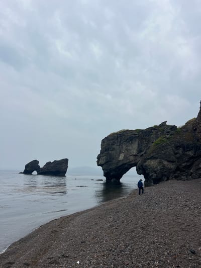 The photograph shows a beach in Gvozdevo, Russia. The sky is overcast. In the center of the frame are two rock formations with natural arches. A person wearing a blue jacket is standing near the base of the larger rock formation. The beach is composed of dark-colored pebbles. The water is reflecting the sky.