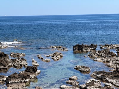 The image depicts a coastal scene in Cala Rajada, Spain. A shallow pool of clear turquoise water is surrounded by dark, porous rocks of varying sizes, some submerged and others exposed. The rocks appear to be of volcanic origin, with a rough texture and a color palette of dark browns and greys. Beyond the pool, the deeper blue waters of the Mediterranean Sea stretch to the horizon under a clear, cloudless sky. A small sailboat can be seen in the distance. The scene suggests a rocky coastline with calm, clear waters, typical of the Mediterranean environment. No people are visible in the image.
