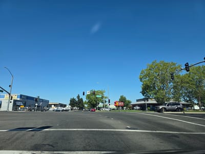 This media file depicts a street-level view of an intersection on a clear, sunny day. The sky is a vibrant, cloudless blue, suggesting it is daytime. The scene is urban, with several commercial buildings visible along the street. On the left, a building with "E-Z SMOG" and "MOG" signs is prominent, along with text indicating services offered, including "WE ALSO DO 'STAR' SHOGS" and mentions of "L& HYBRIDS." Next to it, another building is identified as "ANGNISSCIS Auto Tech Repair."
Further down the street, to the right, a red sign with a starburst shape and the text "Carl's Jr" clearly identifies a fast-food restaurant. Another building, likely associated with the restaurant, has a dark roof. Trees with lush green foliage are scattered throughout the scene, providing shade and adding to the visual landscape. Traffic signals are present at the intersection, and several vehicles, including cars and trucks, are visible on the road, suggesting normal traffic flow. The road itself is asphalt with white lane markings. The overall impression is a typical commercial street intersection in a suburban or urban environment, bathed in bright daylight.