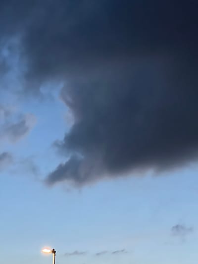 A dark, menacing cloud occupies the majority of the sky in this image, which appears to have been taken in Stoke-on-Trent, United Kingdom. The cloud is a deep, ominous grey, with lighter, wispy edges that suggest movement and the potential for rain. Peeking through a gap in the clouds, a patch of clear, pale blue sky is visible. In the lower portion of the frame, a single streetlamp stands, its bright, warm light cutting through the otherwise somber atmosphere. The lamp is silhouetted against the sky, its post and fixture clearly defined. The overall impression is one of impending weather change, with the lone streetlamp offering a small beacon of light amidst the darkening sky.