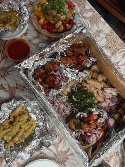 This image captures a generous spread of food, likely a takeout or delivered meal, laid out on a table covered with a cream-colored tablecloth featuring a floral lace pattern. The setting appears to be indoors, possibly for a casual gathering in Zima Yuzhnaya, Russia.

The central focus is a large, foil-lined cardboard box containing an assortment of grilled meats, indicative of shashlik or kebab. This main box is divided into several sections. One section holds cubed, reddish-brown grilled meat, presumably pork or lamb shashlik, generously topped with thinly sliced red onions. Another section contains lighter-colored, elongated pieces of grilled meat, possibly lula kebab or chicken shashlik, also garnished with red onions. A third area features grilled chicken pieces, some appearing to be wings, with their characteristic crispy skin and red onion topping. A large sprig of fresh green herbs, likely cilantro or parsley, is centrally placed, adding color and freshness.

To the left of the main box, a round paper tray holds a vibrant mix of grilled vegetables, including red and yellow bell peppers, mushrooms, and zucchini slices, also garnished with green herbs. A clear plastic cup filled with a reddish-orange sauce, presumably a traditional shashlik sauce, is visible near the bottom center.

Further down, two portions of what appears to be mashed potatoes or a similar starchy side dish are individually wrapped in aluminum foil, suggesting they are meant to be kept warm. One is on a small white plate on the bottom left, and another is towards the bottom right.

The overall scene suggests a hearty and celebratory meal, prepared for sharing, characterized by its varied grilled offerings and the practical, disposable packaging. No identifiable text is clearly visible on any items.