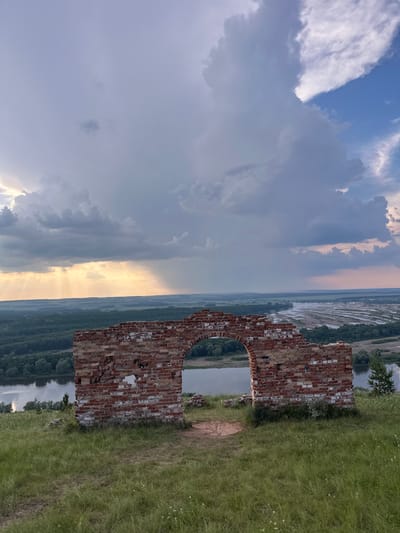 This is a photograph taken from a grassy hilltop overlooking a wide river valley. In the foreground, a ruined brick archway stands prominently. The archway is made of weathered red bricks with some white mortar visible, and it has a large opening in its center. The surrounding ground is covered in green grass with a few small white wildflowers.
Beyond the archway, the landscape unfolds with a broad river winding through a verdant valley. The river appears to have multiple sandbars or shallow areas. The valley is densely forested with various shades of green trees. In the distance, the land flattens out, and a hazy horizon is visible under a dramatic sky.
The sky is a mix of stormy gray clouds and patches of brighter blue. Sunlight is breaking through the clouds in some areas, casting visible rays of light downwards, particularly on the left side of the image, illuminating the distant landscape and river. The overall mood is one of natural beauty and historical remnants.
The location is identified as Nizhniye Vyazovye, Russia.