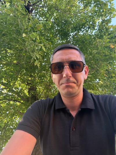 A man is taking a selfie outdoors, with a tree and its green leaves filling the background. The man, with short grey hair, is wearing a black polo shirt and sunglasses. He is looking directly at the camera with a neutral expression. The lighting suggests it is daytime. The scene takes place in Parma, Italy, in an outdoor setting.